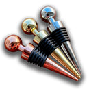 Stoppers metálicos Johnny Cooker nas cores cobre, dourado e prata, exibindo acabamento de alto brilho e anéis de silicone pretos para vedação hermética de garrafas de vinho.