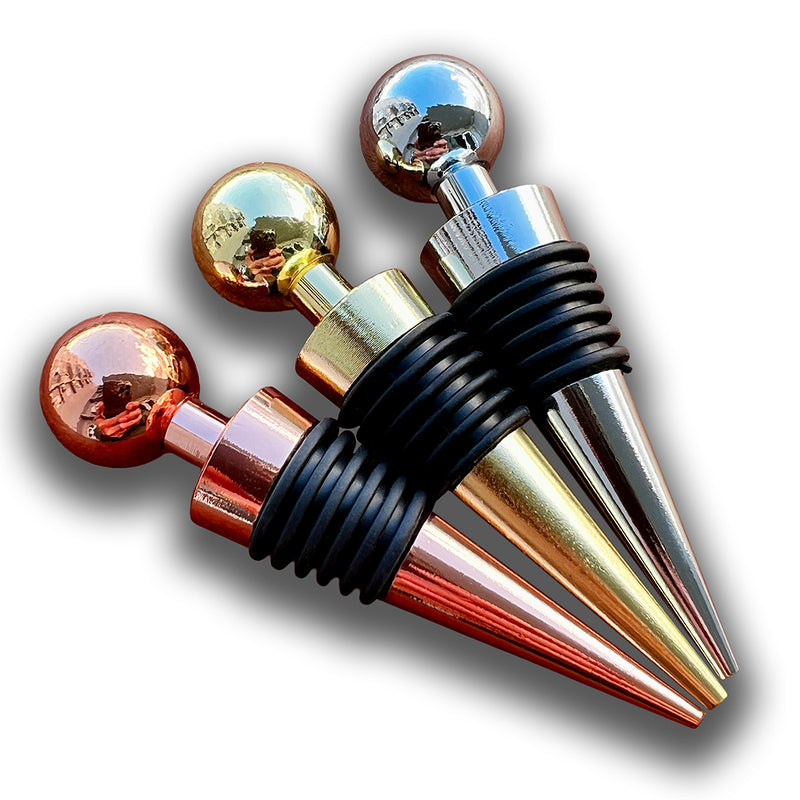 Stoppers metálicos Johnny Cooker nas cores cobre, dourado e prata, exibindo acabamento de alto brilho e anéis de silicone pretos para vedação hermética de garrafas de vinho.