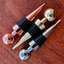 Stoppers metálicos Johnny Cooker nas cores cobre, dourado e prata, exibindo acabamento de alto brilho e anéis de silicone pretos para vedação hermética de garrafas de vinho.