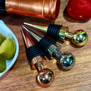 Stoppers metálicos Johnny Cooker nas cores cobre, dourado e prata, exibindo acabamento de alto brilho e anéis de silicone pretos para vedação hermética de garrafas de vinho.