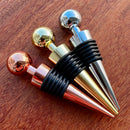 Stoppers metálicos Johnny Cooker nas cores cobre, dourado e prata, exibindo acabamento de alto brilho e anéis de silicone pretos para vedação hermética de garrafas de vinho.