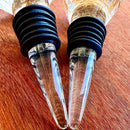 Stopper Cristal Gold Johnny Cooker para garrafas de vinho, com haste cônica , equipadas com anéis de vedação de silicone preto. Topos em material transparente facetado (nos formatos diamante e gota), revelando em seu interior um preenchimento denso de folhas douradas brilhantes.