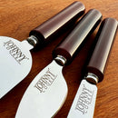 Três facas para queijos da marca Johnny Cooker com lâminas de aço inox polido (modelos lança, cutelo e espátula) e cabos cilíndricos em pedra natural Ágata polidos brilhantes.