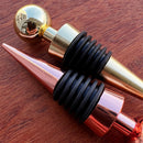 Stoppers metálicos Johnny Cooker nas cores cobre, dourado e prata, exibindo acabamento de alto brilho e anéis de silicone pretos para vedação hermética de garrafas de vinho.