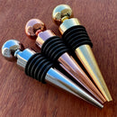 Stoppers metálicos Johnny Cooker nas cores cobre, dourado e prata, exibindo acabamento de alto brilho e anéis de silicone pretos para vedação hermética de garrafas de vinho.