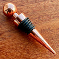 Stoppers metálicos Johnny Cooker nas cores cobre, dourado e prata, exibindo acabamento de alto brilho e anéis de silicone pretos para vedação hermética de garrafas de vinho.